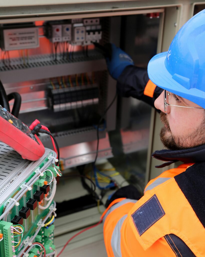 Electrician wearing safety gear operates switchboard in industrial setting.